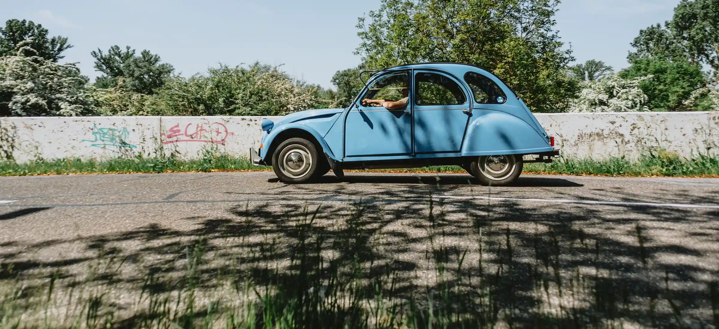Une voiture ancienne arrive sur une route de campagne au pied des Pyrénées, vue depuis le bas-côté